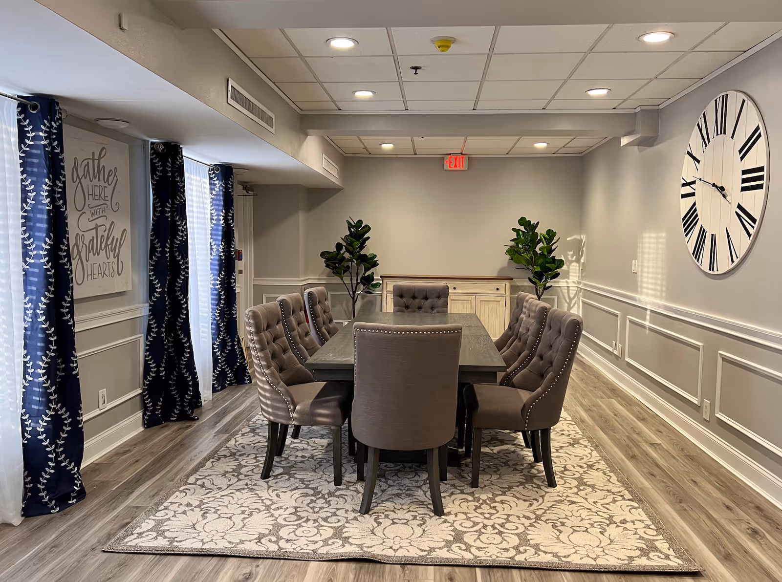 A dining room with a rectangular wooden table surrounded by eight upholstered chairs. The room has light gray walls with white wainscoting, a large decorative wall clock with Roman numerals on the right wall, and two tall green plants flanking a wooden sideboard at the back. Blue curtains with white leaf patterns hang over windows on the left side, and a wall sign reads 'gather here with grateful hearts'. The floor is covered with a patterned area rug beneath the table and chairs.