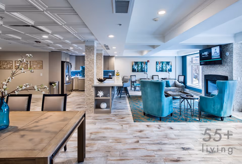 A bright and modern senior living common area featuring a wooden dining table with chairs and a blue vase with white flowers, a kitchen with stainless steel appliances and a breakfast bar with stools, and a cozy living room with teal armchairs, a sofa, a fireplace, and wall art. The space has light wood flooring and recessed lighting.