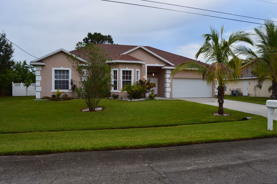 Single-story suburban house with a two-car garage, manicured lawn, and palm trees under an overcast sky.