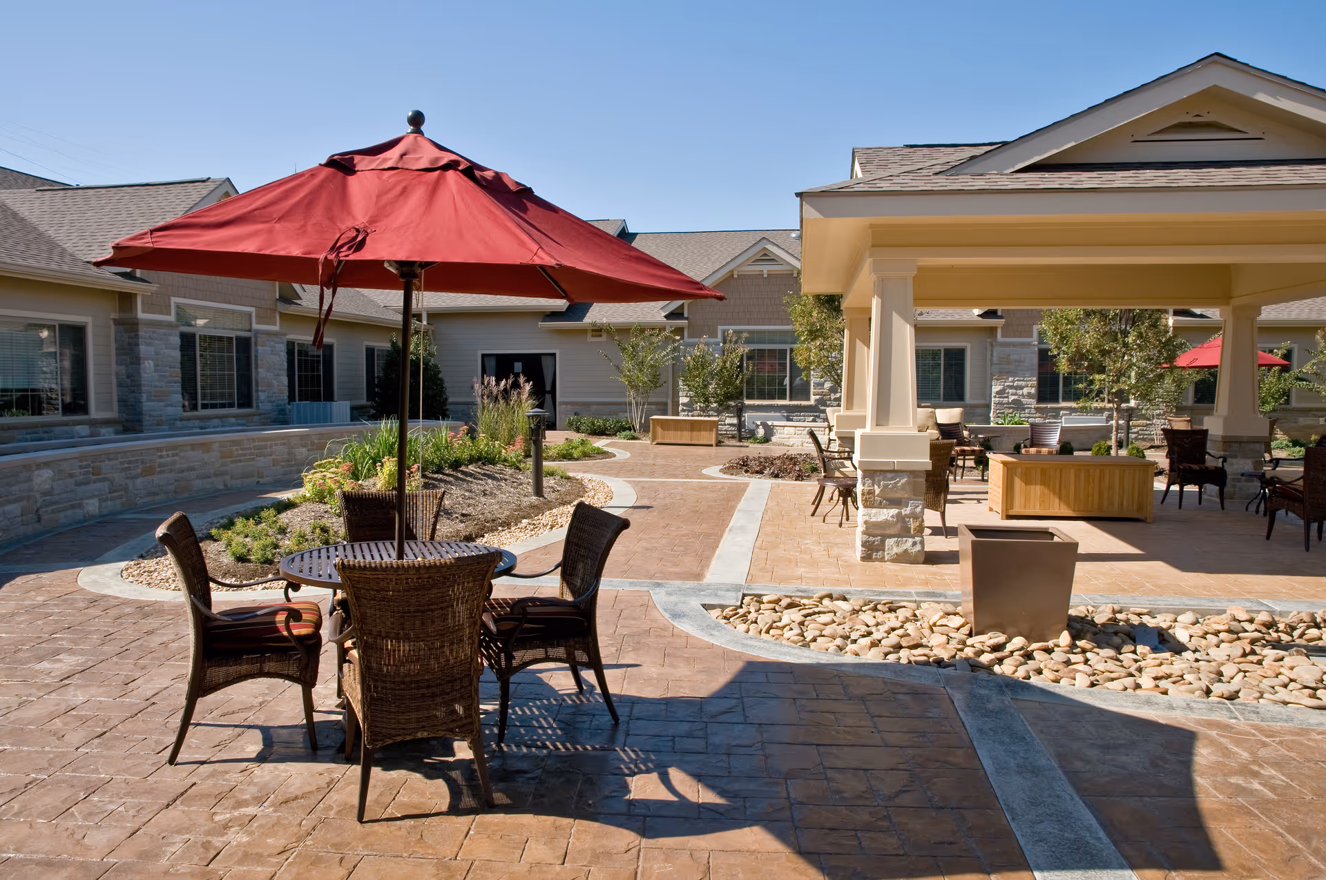 Outdoor patio area at a senior living facility with a round table and four wicker chairs under a large red umbrella. The patio is paved with stone tiles and features landscaped garden beds with plants and small trees. There is a covered seating area with additional chairs and tables in the background, along with a building exterior with windows.