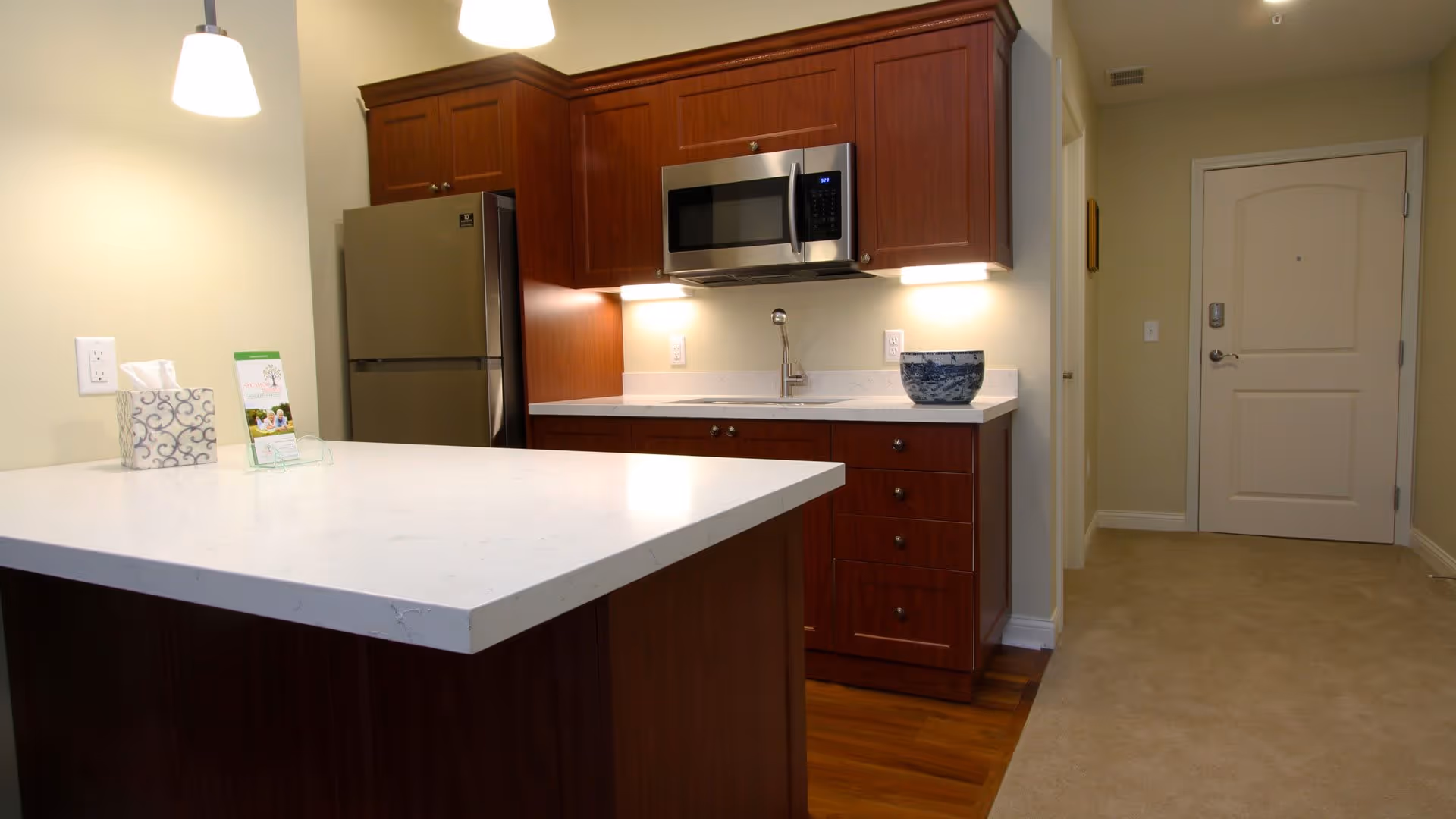 Modern kitchen area in a senior living facility with wooden cabinets, a stainless steel refrigerator, a microwave, a sink, and a white countertop island. The kitchen is well-lit with pendant lights and under-cabinet lighting. A hallway with a closed door is visible to the right.