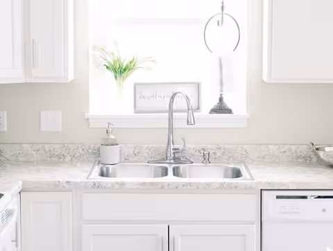 Bright kitchen sink area with a double stainless steel sink, a modern curved faucet, white cabinetry, and a marble-patterned countertop. A window above the sink lets in natural light and has a small plant and decorative items on the sill.