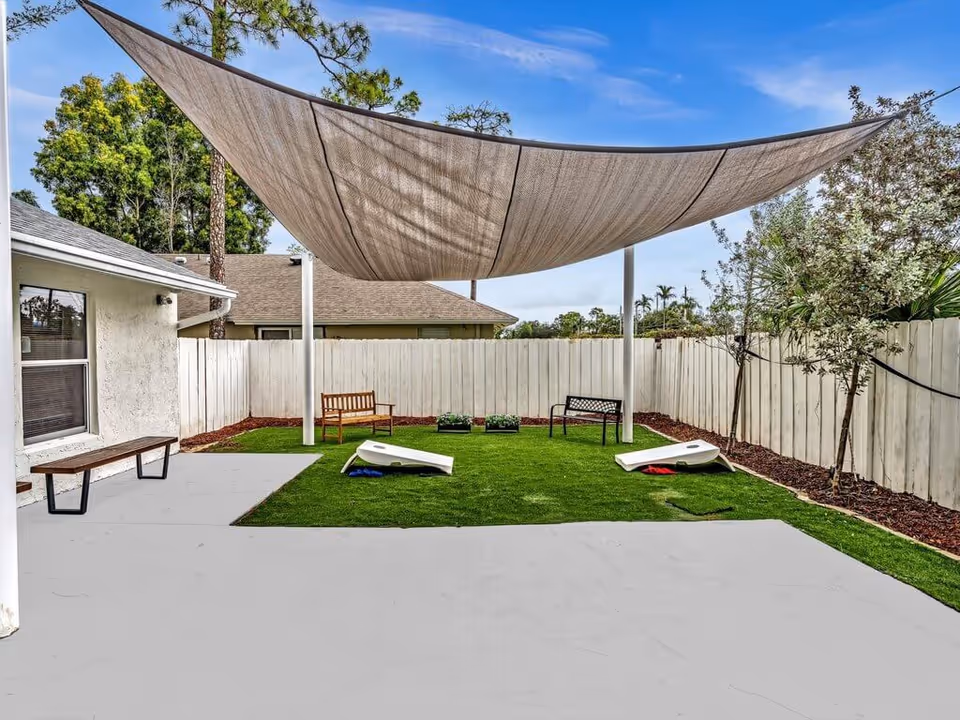Fenced backyard patio with a large triangular shade sail, benches, artificial turf and cornhole boards.