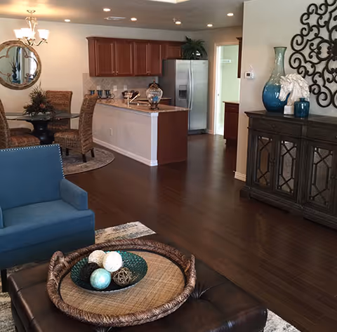 Interior view of a senior living facility showing a living room area with a blue armchair and a dark ottoman with a decorative tray on top. In the background, there is a dining area with a round glass table and wicker chairs, and a kitchen with wooden cabinets and stainless steel refrigerator. Decorative items and wall art are visible on the right side.