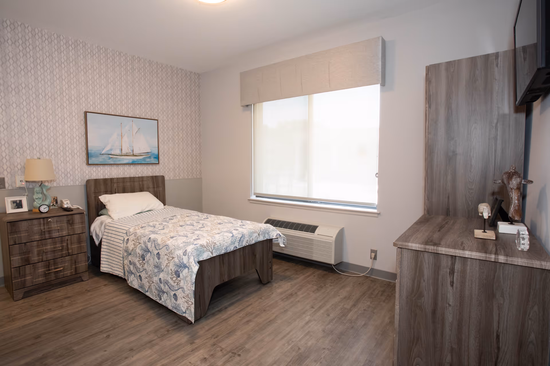 A single bedroom in a senior living facility with a wooden bed featuring a floral bedspread and a pillow. Next to the bed is a wooden nightstand with a lamp, clock, phone, and framed photo. The room has a large window with a beige valance and a white roller shade, and a wooden dresser with decorative items on top. The floor is wood laminate, and the walls are painted light gray with a patterned wallpaper accent behind the bed.
