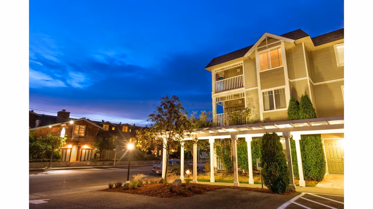 Dusk exterior view of a multi-story retirement inn with illuminated entrance, covered walkway, landscaping, and adjacent street.