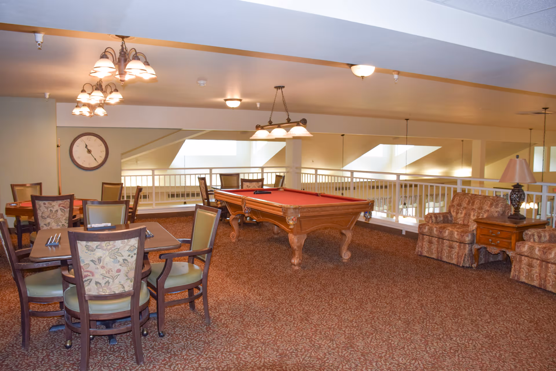 Interior view of a senior living facility common area featuring a red pool table in the center, several tables with chairs around them, and a seating area with two patterned armchairs and a wooden side table with a lamp. The space has carpeted flooring, multiple ceiling lights, and a large clock on the wall.