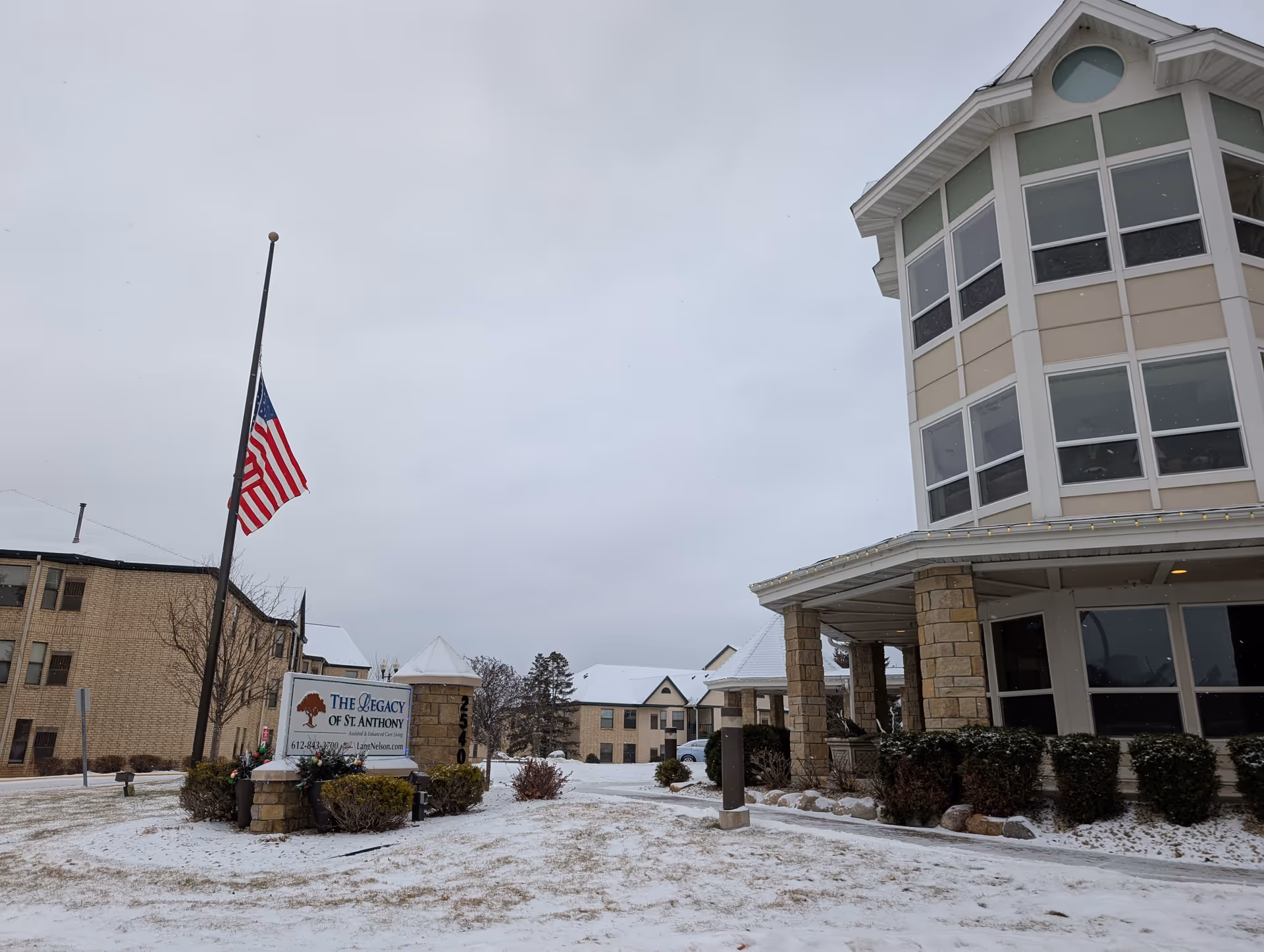 Exterior view of a senior living facility with snow-covered ground and buildings. A flagpole with an American flag at half-mast stands next to a sign that reads 'The Legacy of St. Anthony Assisted & Enhanced Care Living'. The buildings have beige brick walls and large windows, with some greenery around the entrance.