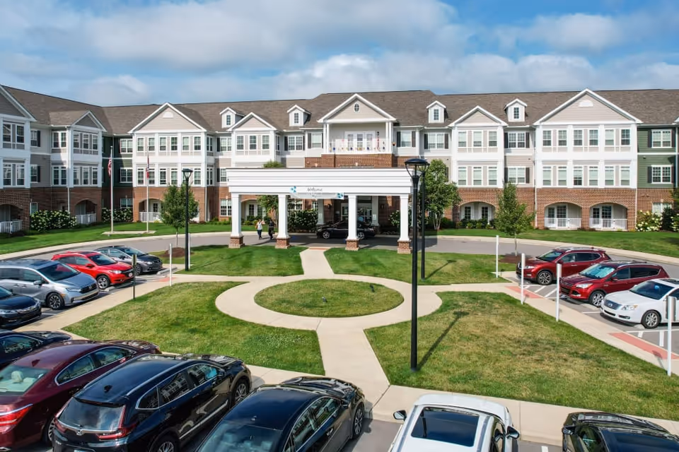 Front exterior view of a large senior living facility named Harmony at Victory Station with a circular driveway, parked cars, green lawns, and a clear blue sky.