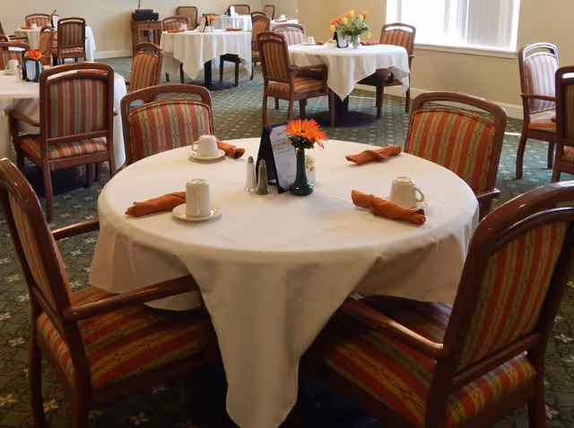 Round dining table set with a white tablecloth, cups, orange napkins, and a flower centerpiece in a dining room with additional similarly set tables.