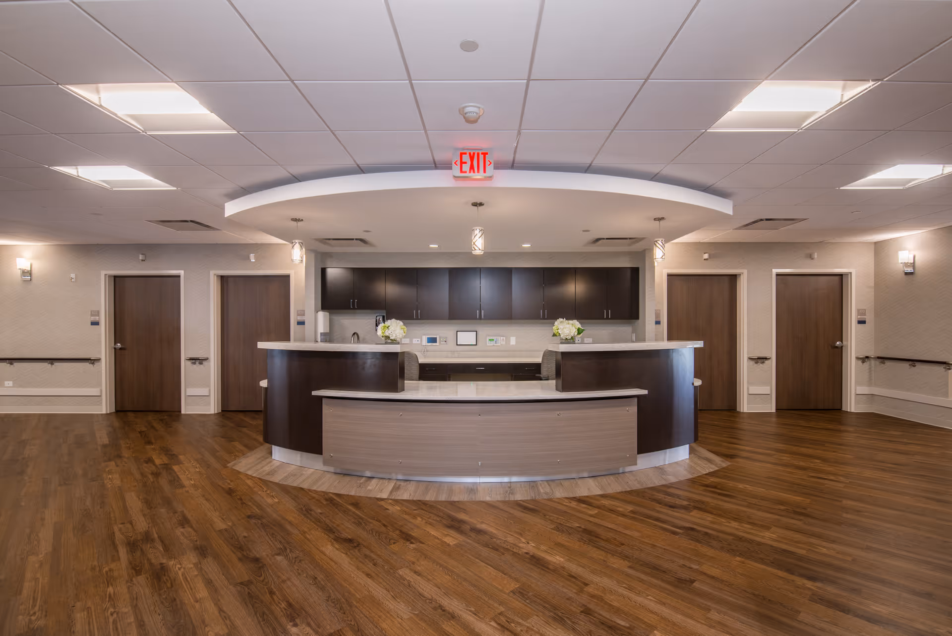 A modern reception desk area in a senior living facility with a curved counter, dark wood cabinetry, and light wood flooring. The ceiling has recessed lighting and hanging pendant lights. There are four closed wooden doors visible in the background, and an illuminated red exit sign is mounted above the reception area.