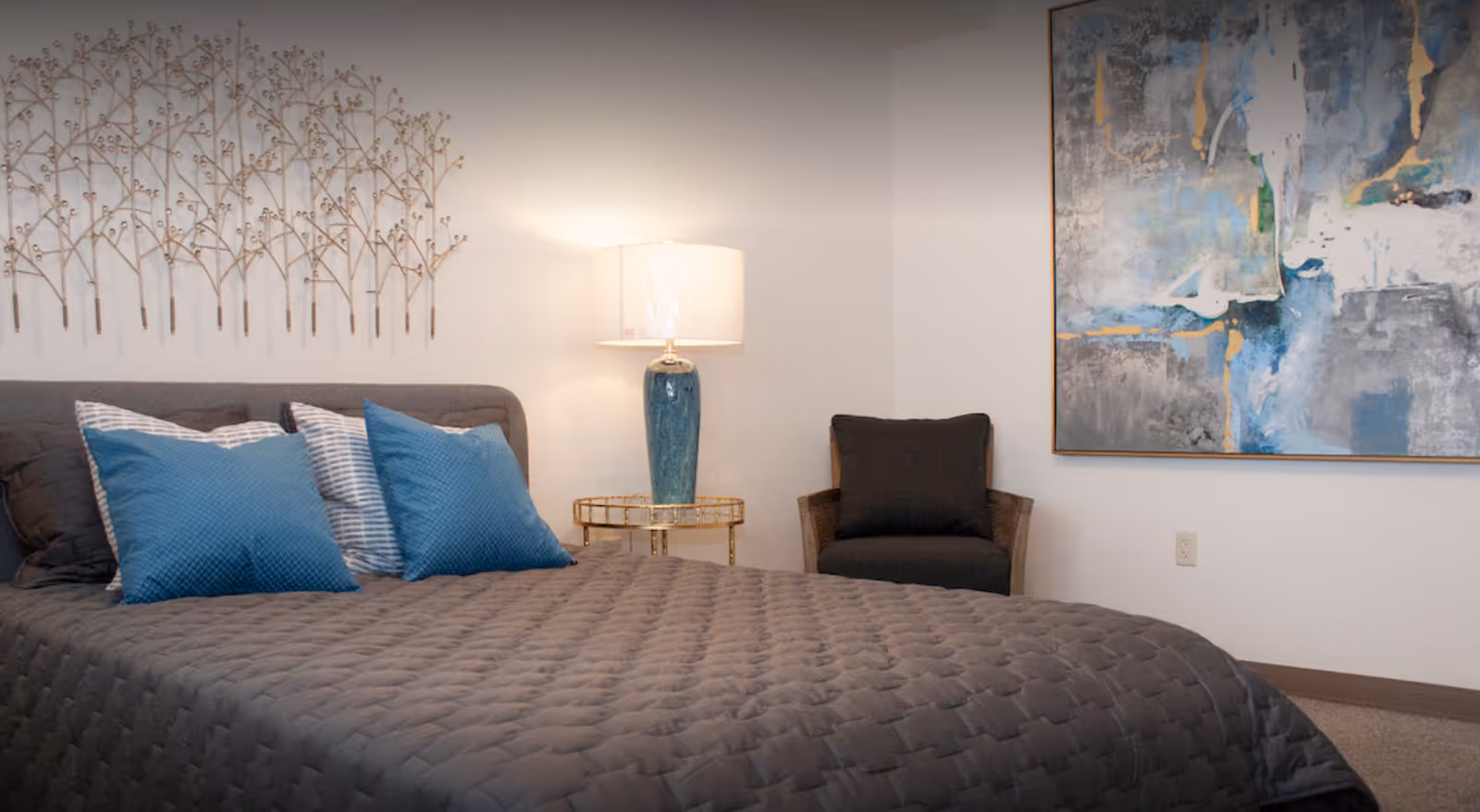 A bedroom with a gray quilted bedspread and four pillows, two blue and two striped. There is a blue ceramic table lamp on a gold side table next to the bed. A dark cushioned chair is placed in the corner, and a large abstract painting with blue, gray, and gold tones hangs on the wall. Decorative metal wall art resembling tree branches is mounted above the bed.