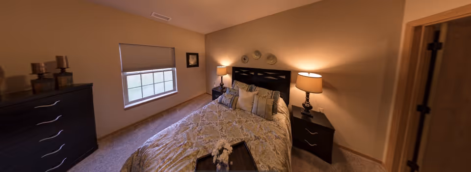 A cozy bedroom with a bed covered in patterned bedding and multiple pillows, flanked by two nightstands each with a lamp. There is a window with a blind partially closed on the left wall, a dresser with decorative items on top, and a door on the right side of the room. The walls are painted beige and the floor is carpeted.