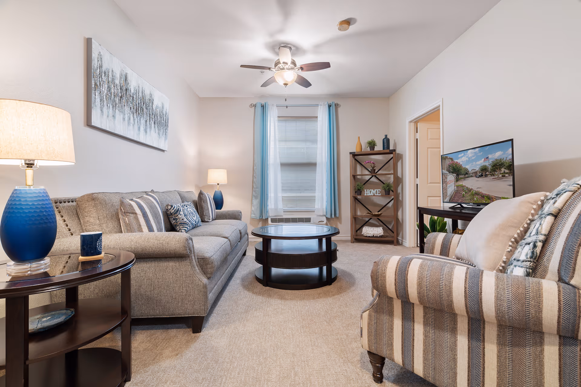 A furnished living room featuring a gray sofa, striped armchair, round coffee table, TV and decorative shelving with blue accents.