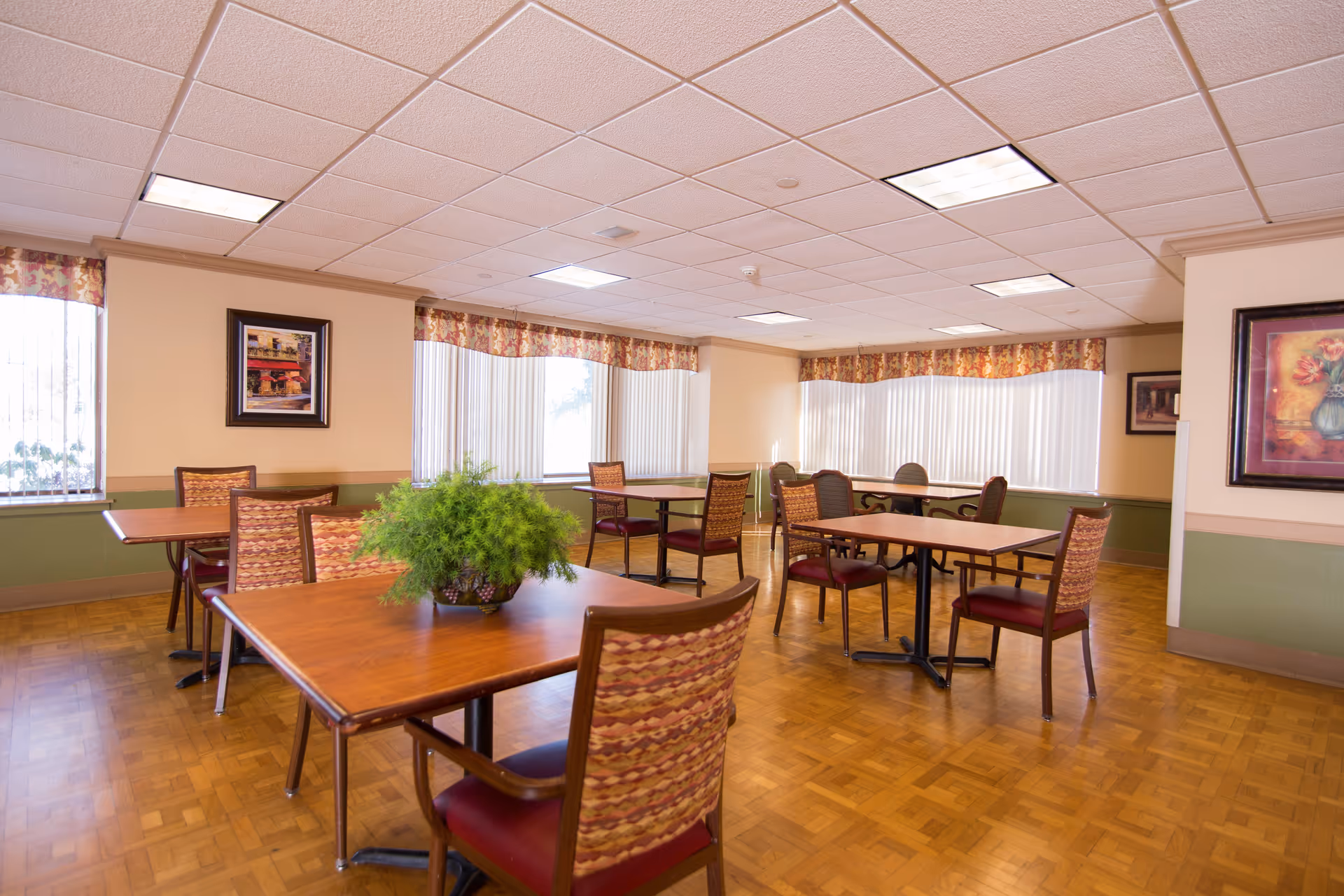 A bright dining room with several wooden tables and chairs arranged neatly. The room has large windows with vertical blinds and floral valances, parquet flooring, and framed artwork on the walls. A green potted plant is placed on one of the tables.