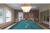 A well-lit room with a green pool table in the center, featuring a cue stick and a white cue ball on the table. The room has large windows on the left side letting in natural light, a hanging light fixture above the pool table, and seating with chairs and a small table against the far wall. There is a framed picture on the wall and a rack holding pool cues on the right side.