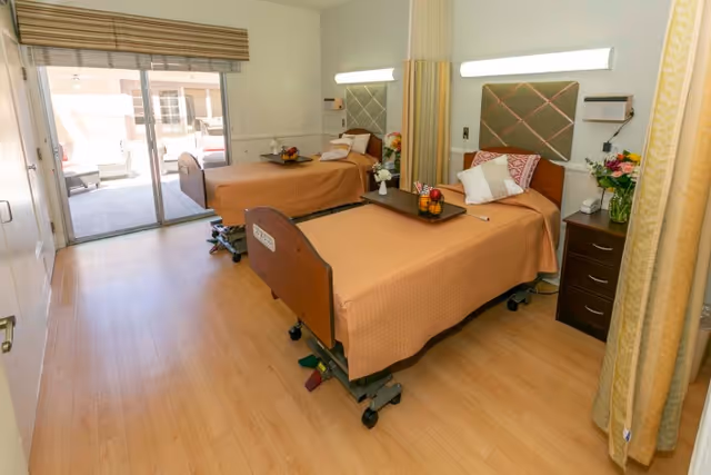 A bright room with two hospital-style beds covered with light brown bedspreads, each with pillows and a tray holding fruit. The room has light wood flooring, a sliding glass door leading to an outdoor patio area, and a curtain divider between the beds. There are nightstands with flowers and a wall-mounted light above each bed.