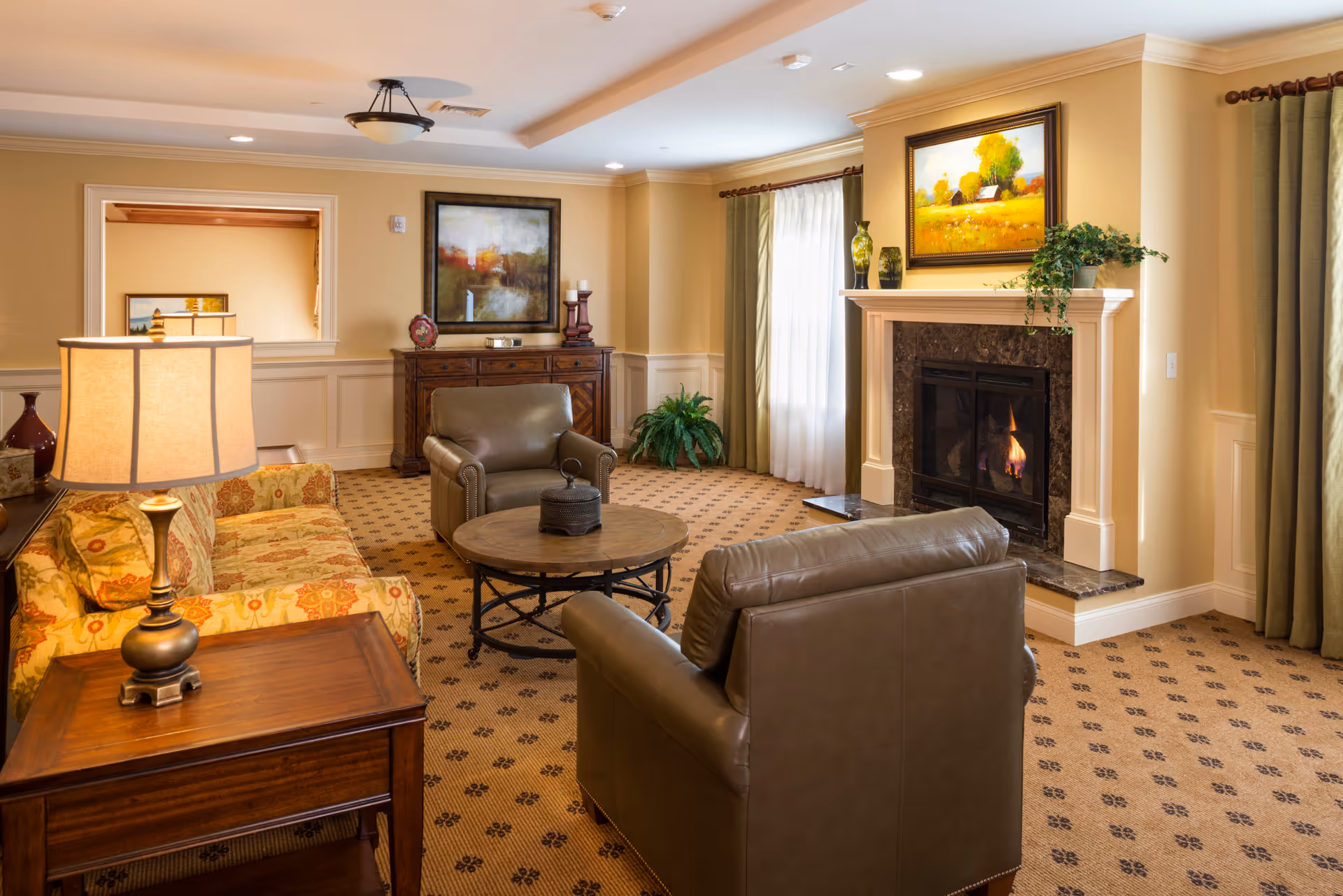 Cozy furnished living room with chairs, a sofa, coffee table and a lit fireplace under framed artwork.