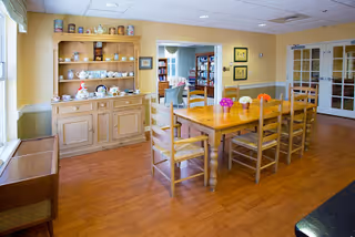 A dining room with a wooden table and six chairs. On the table are small flower arrangements. Against the wall is a wooden hutch displaying various teapots and dishes. The room has light yellow walls, wood flooring, and a doorway leading to another room with bookshelves.
