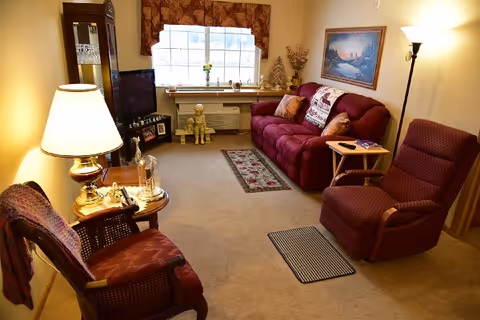 A cozy living room in an assisted living facility featuring a burgundy sofa with pillows, a matching armchair, a wooden chair with a cushion, a floor lamp, a TV on a stand, a window with floral curtains, and decorative items including a small statue and framed pictures.