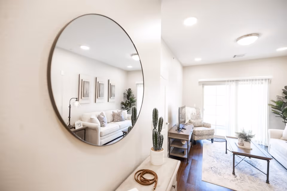 Bright and airy living room with a white sofa, patterned armchair, wooden coffee table, and a TV on a stand. A large round mirror on the wall reflects part of the sofa and framed artwork. The room has hardwood floors, a large window with sheer curtains, and potted plants adding greenery.