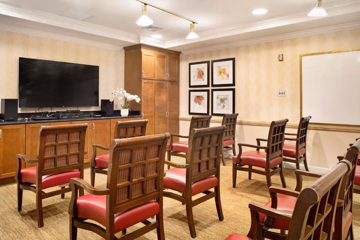 A small meeting or activity room with wooden chairs featuring red cushions arranged in rows facing a large flat-screen TV mounted on a wooden cabinet. The room has beige patterned wallpaper, framed artwork on the wall, a whiteboard, and ceiling lights.