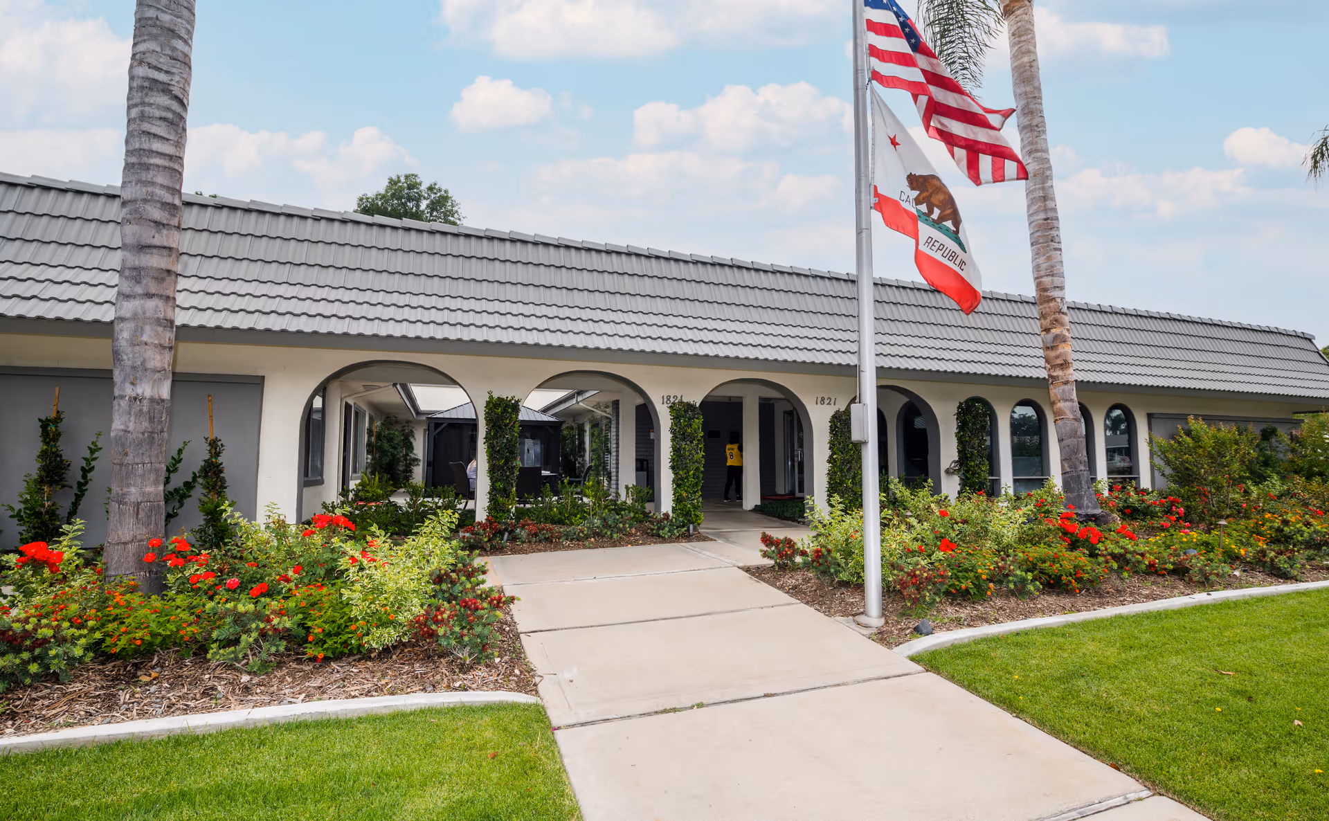 Front exterior view of Gordon Lane Care Center building with a tiled roof, arched entryways, and a landscaped garden with red flowers and palm trees. Two flagpoles display the American flag and the California state flag.