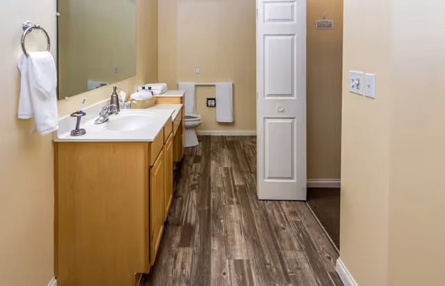 A clean bathroom with a wooden vanity and sink, large mirror, toilet, towel rack, and wood-look flooring.