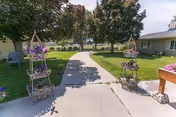 Outdoor paved walkway flanked by decorative plant stands with purple flowers, leading through a grassy area with large trees and buildings on either side under a partly cloudy sky.