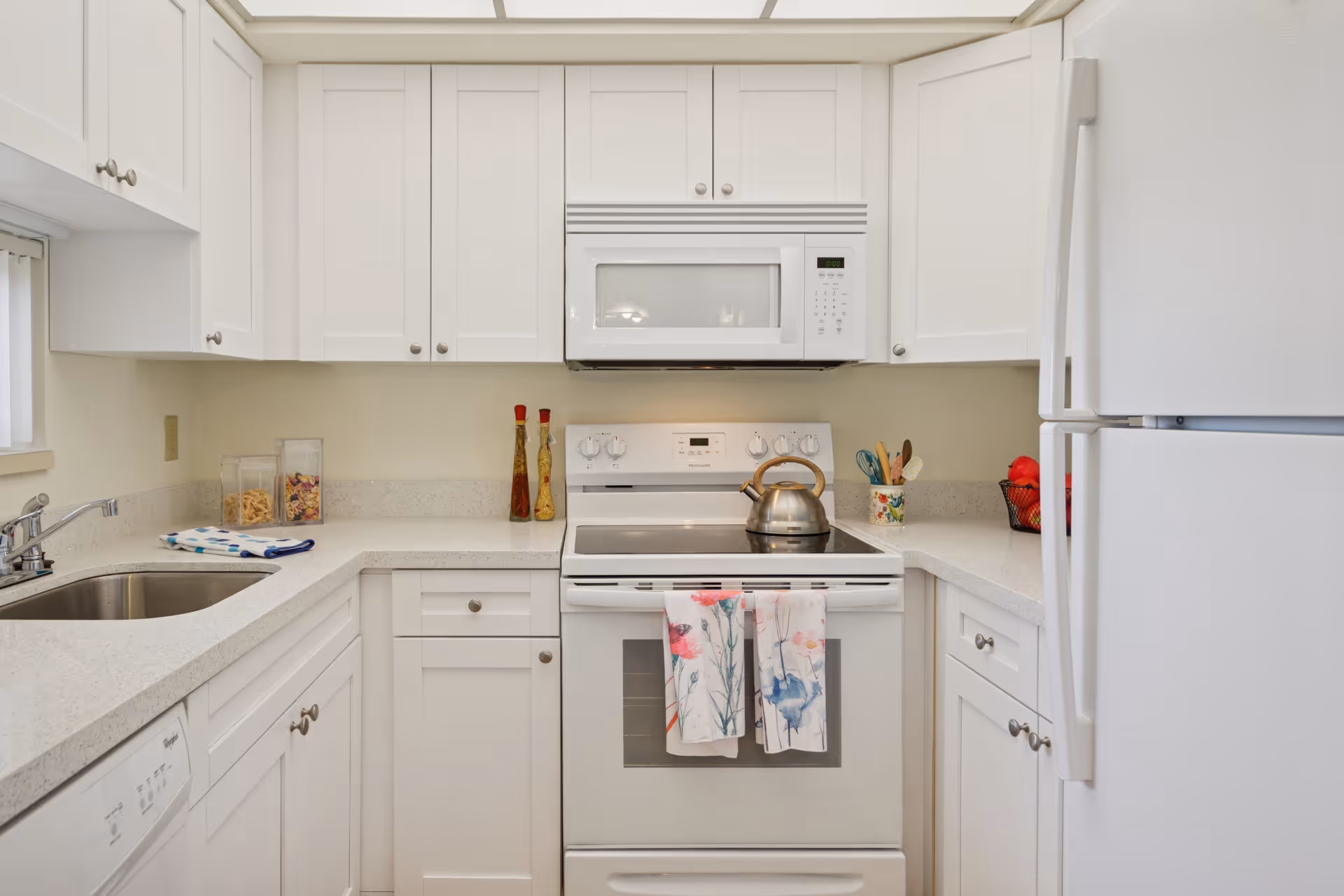 A clean, white kitchen with white cabinets, a white refrigerator, a white stove with a kettle on it, and a white microwave above the stove. There are two colorful dish towels hanging on the oven door, a sink with a faucet on the left side, and various kitchen utensils and containers on the countertops.