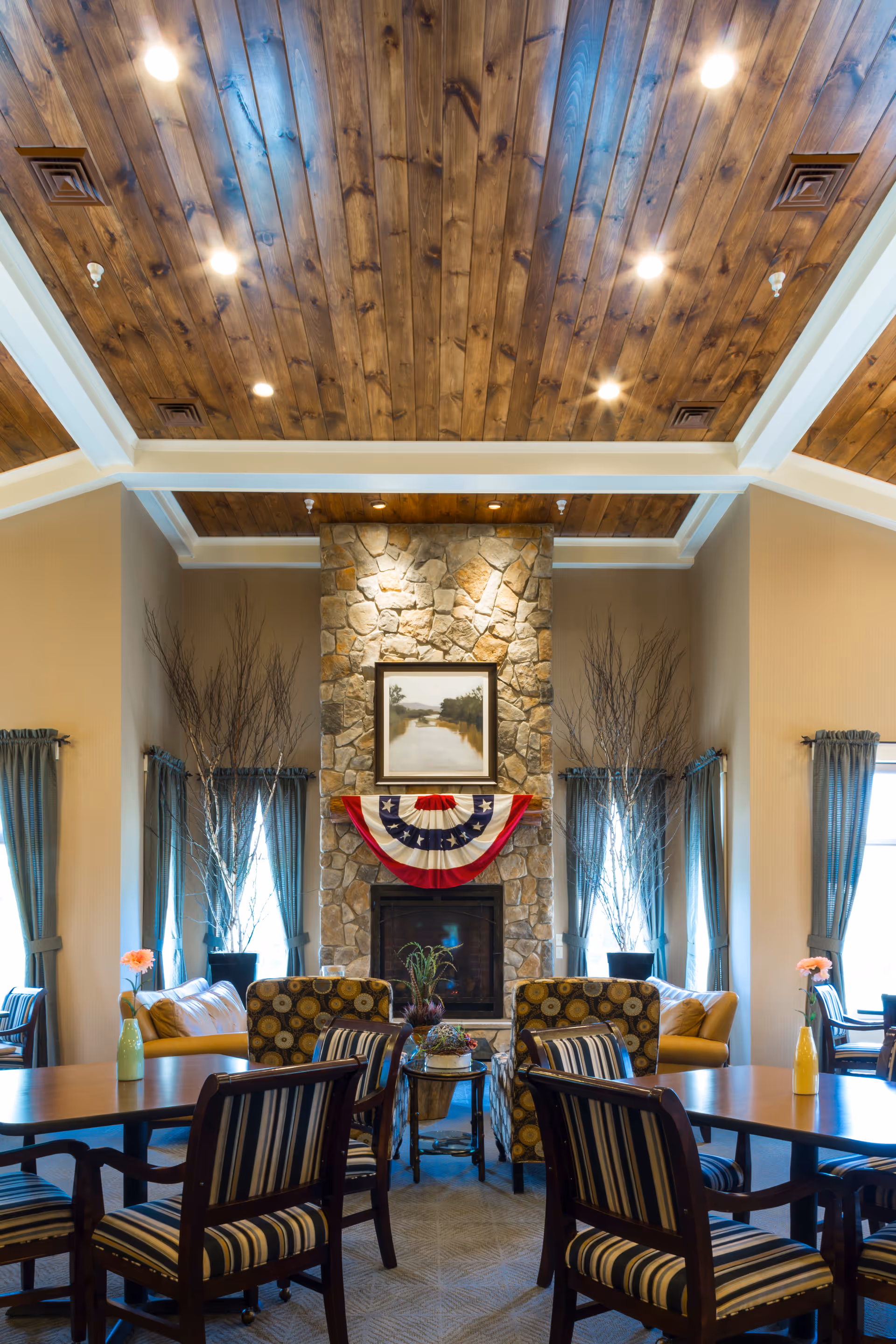 Cozy common room with a stone fireplace, seating, and tables under a wood-paneled ceiling.