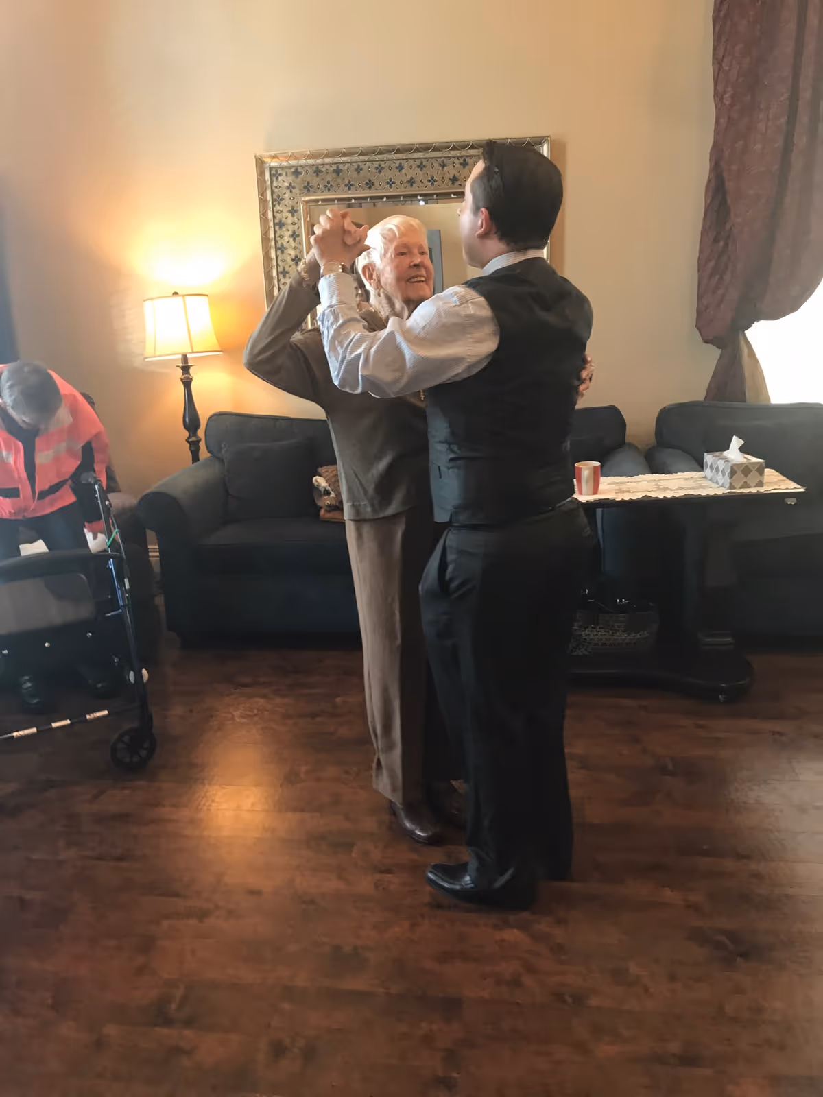 An elderly woman and a man in formal attire are dancing together in a cozy living room with wooden floors. In the background, another elderly person with a walker is seen near a dark-colored couch, a standing lamp, and a table with a tissue box and a cup. The room has warm lighting and a large decorative mirror on the wall.