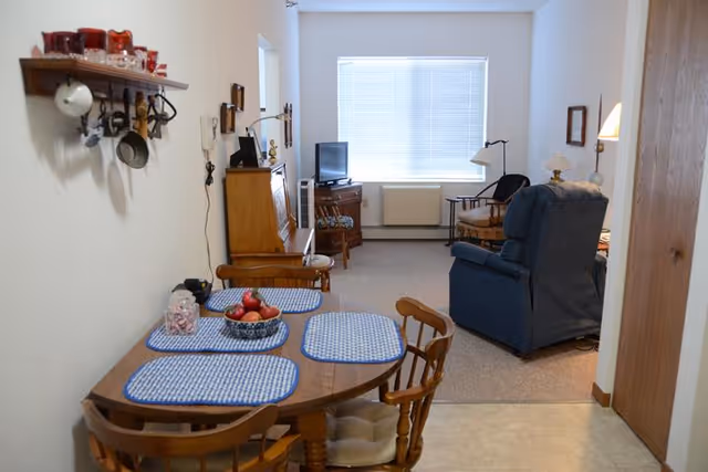 Small assisted-living apartment interior showing a dining table with placemats in the foreground and a living area with a recliner, TV cabinet, and window in the background.