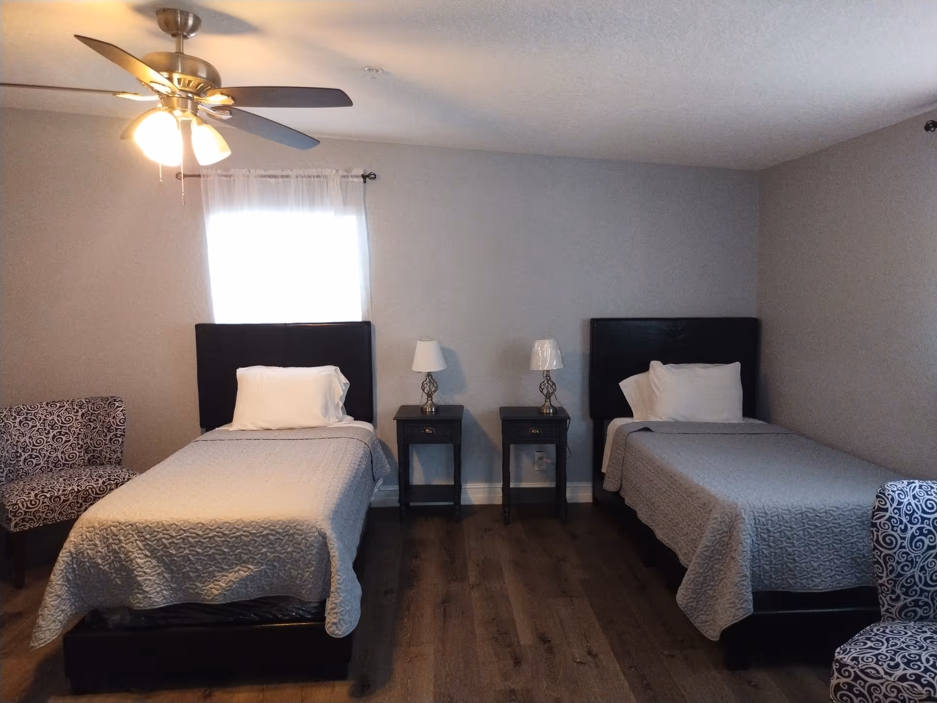 A bedroom with two single beds each with a black headboard, white pillow, and gray quilted bedspread. Between the beds are two small black nightstands, each with a lamp. There are two patterned chairs, one on each side of the beds. A ceiling fan with lights is on the ceiling, and a window with sheer white curtains is behind the left bed.