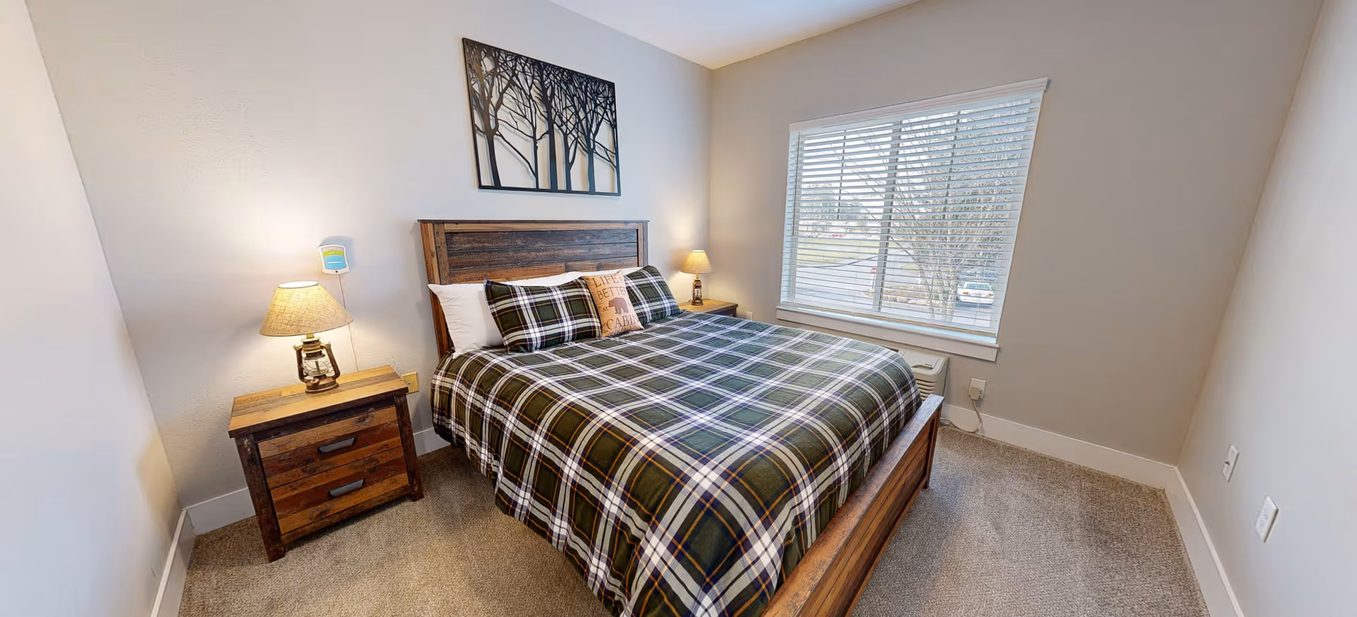 A cozy bedroom with a wooden bed frame and matching nightstands on either side. The bed is made with a green and white plaid comforter and several pillows, including one with the text 'Live Sweet Care'. There are two lamps on the nightstands, and a window with white blinds lets in natural light. A decorative metal wall art piece featuring tree silhouettes hangs above the bed.