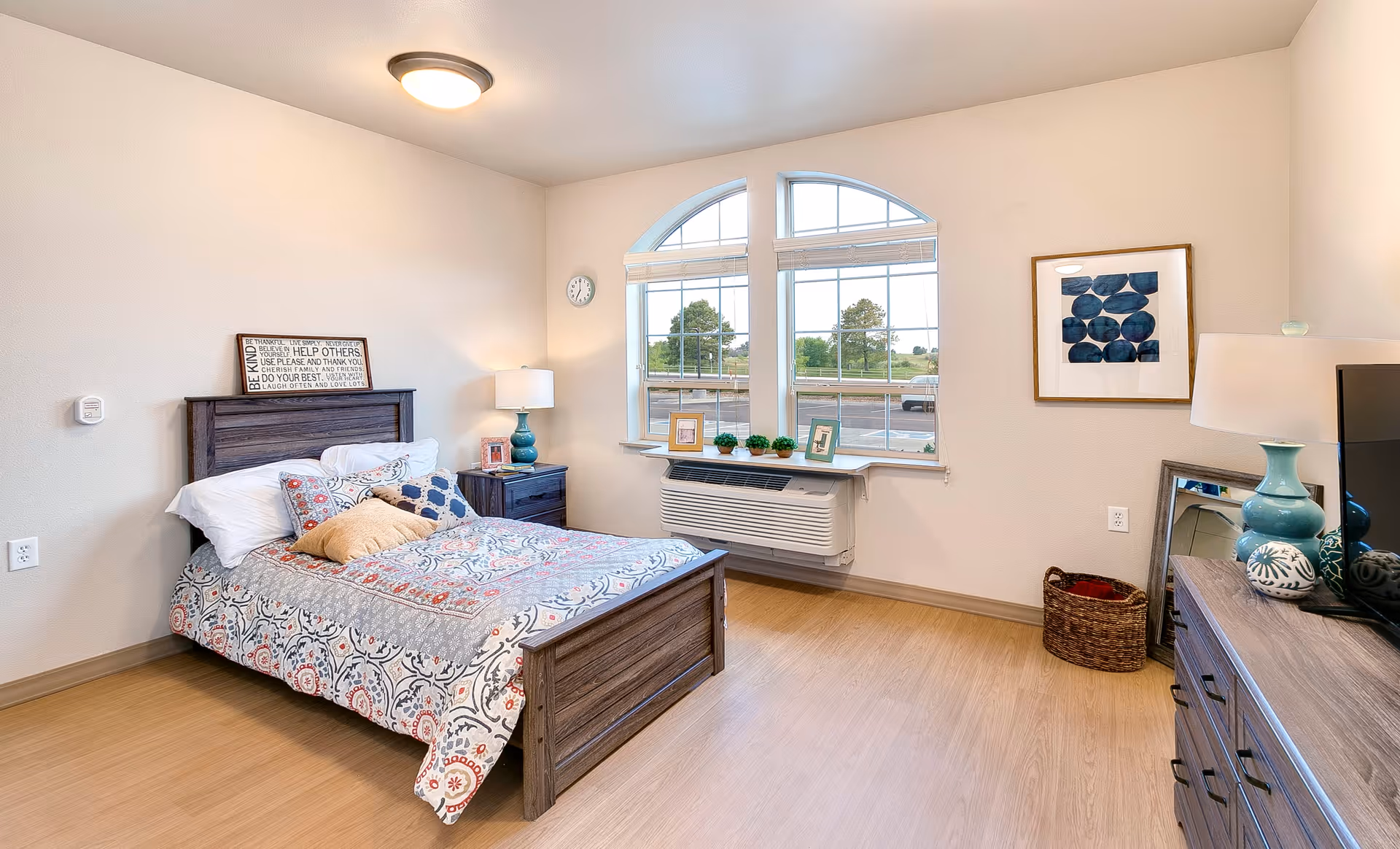 A bright and cozy bedroom in a senior living facility featuring a wooden bed with patterned bedding and multiple pillows. There is a nightstand with a lamp and picture frame beside the bed. The room has large windows with an arched design letting in natural light, a wall clock, a framed abstract artwork, a wicker basket, and a dresser with a lamp and decorative items. The floor is wooden and the walls are painted light beige.