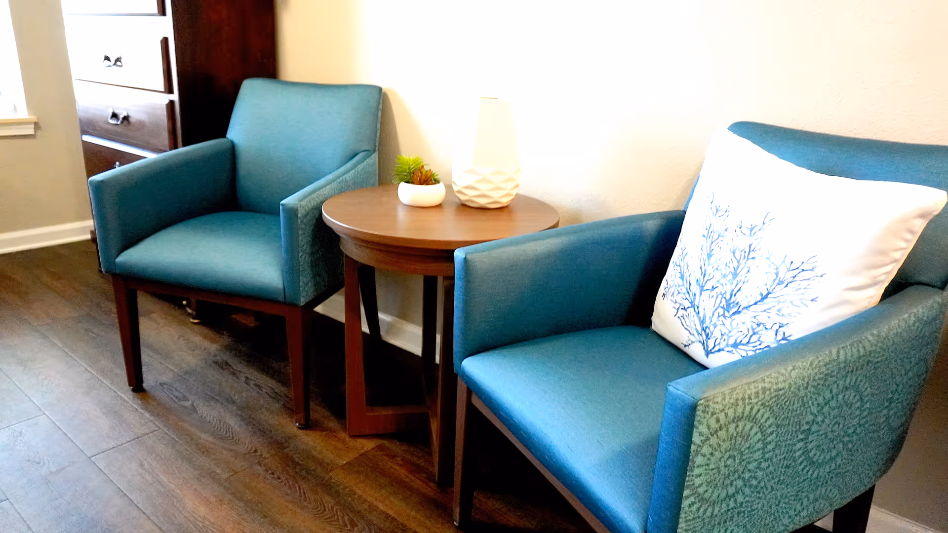 Two teal upholstered armchairs with wooden legs are positioned on either side of a round wooden side table. The table holds a white decorative lamp and a small potted plant. One chair has a white pillow with a blue coral design. The room has wooden flooring and a dresser is visible in the background.