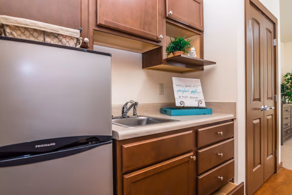 A small kitchenette area with a stainless steel Frigidaire mini refrigerator, a sink with a faucet, wooden cabinets above and below the countertop, and a decorative sign on a stand that reads 'every good and perfect gift is from above'. There is also a small potted plant on a shelf above the countertop. A wooden door and part of another room with a dresser and plant are visible in the background.