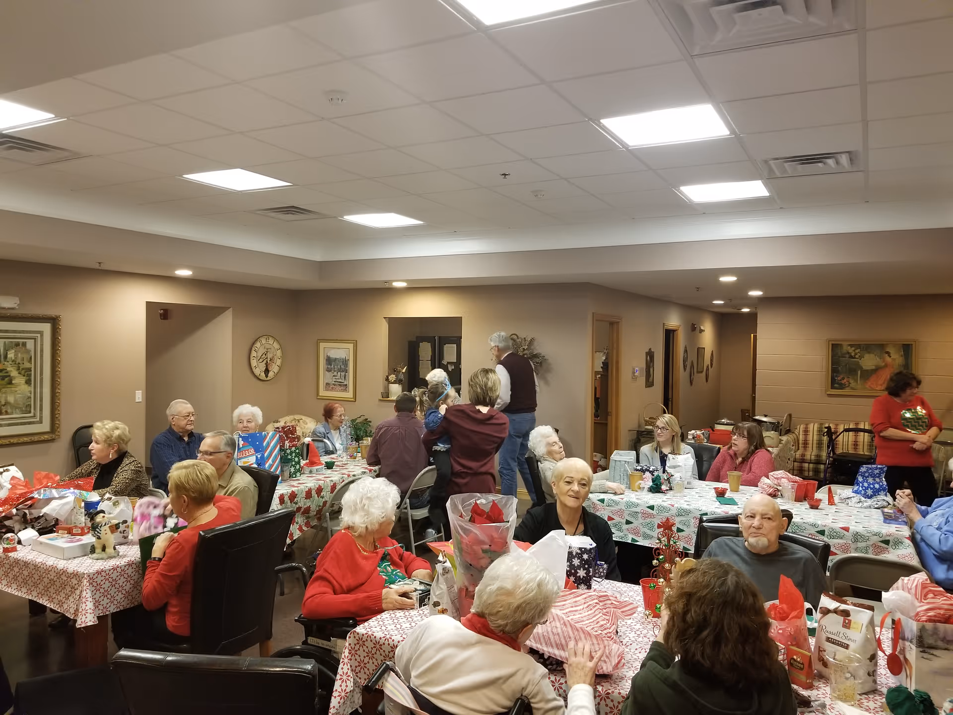 A group of elderly people gathered in a decorated common room, sitting around tables covered with festive holiday tablecloths and surrounded by wrapped gifts and holiday decorations. The room has beige walls, framed pictures, and bright ceiling lights.