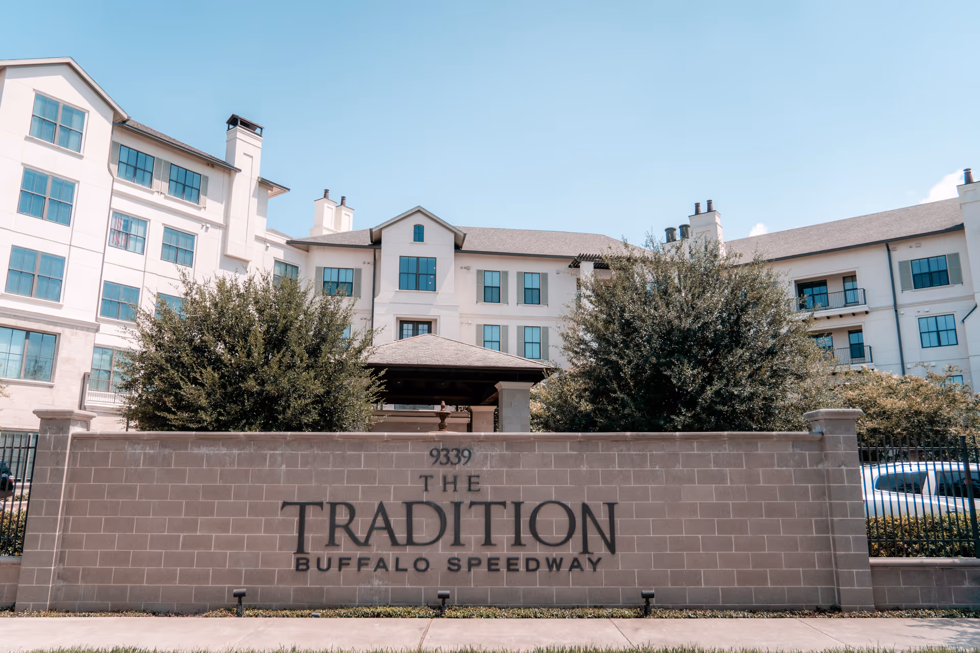 Exterior view of The Tradition Buffalo Speedway building with its name on a brick entrance wall and landscaped shrubs.