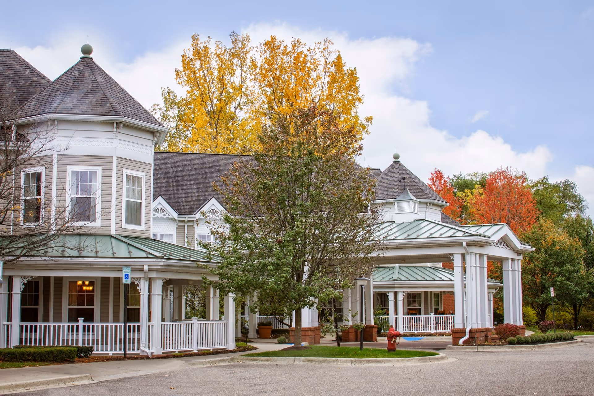 Exterior view of a senior living facility building with a covered entrance and porch area. The building features a turret-style roof and white railings. Trees with autumn foliage in yellow, orange, and red surround the building under a partly cloudy sky.