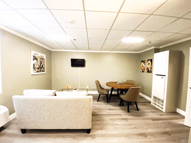 A senior living facility common area with a beige sofa facing a wall-mounted flat screen TV. To the right, there is a round wooden table with four cushioned chairs. The room has light-colored walls, wood flooring, and ceiling tiles. There are two framed artworks on the walls and a white cabinet with cubby holes on the right side.