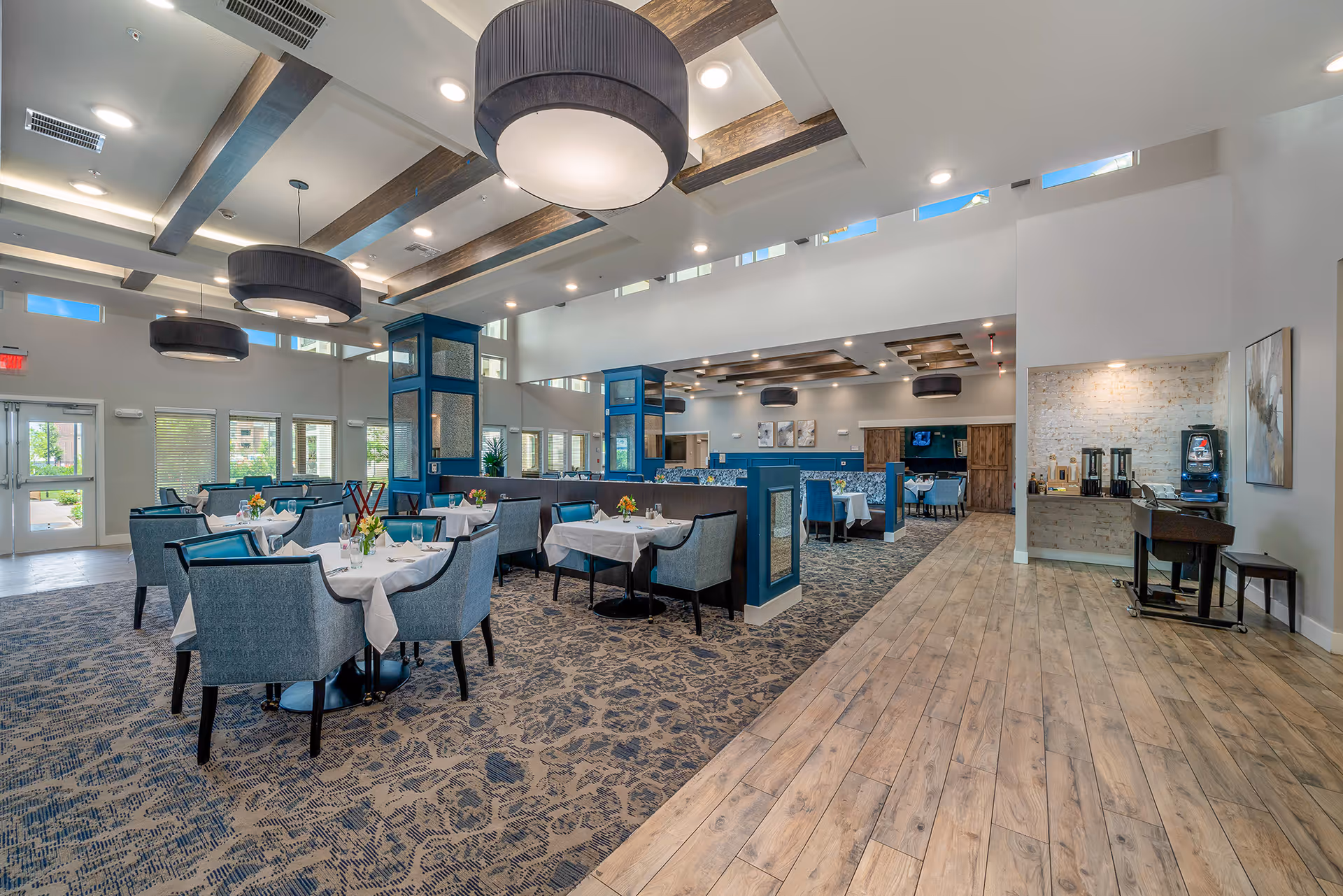 Spacious, well-lit dining room with round tables set with white linens, blue upholstered chairs, large overhead drum lights, and wood-accented ceilings.
