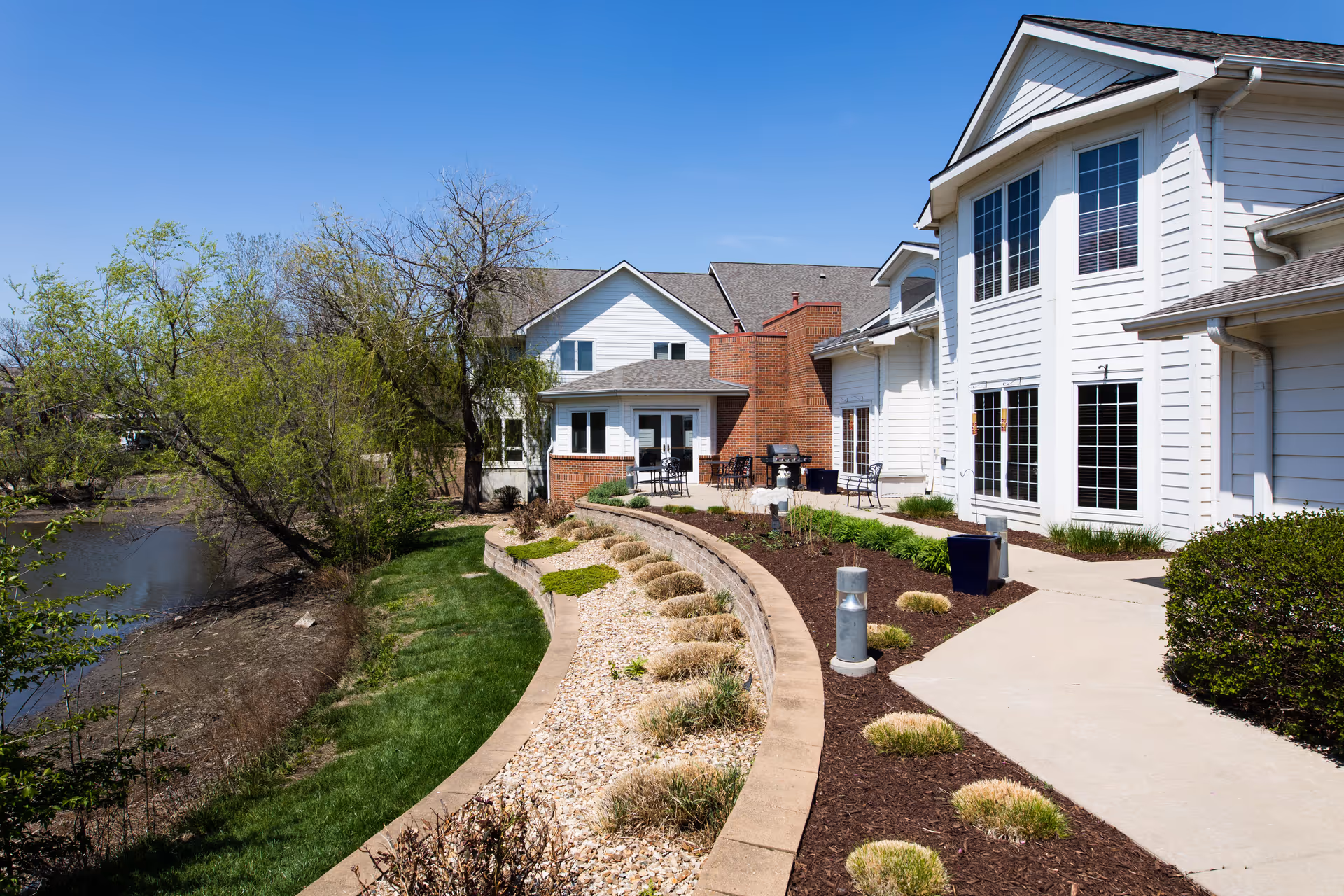 Exterior view of Lexington Park Assisted Living of Topeka showing a white multi-story building with large windows, a landscaped garden with shrubs and grass, a paved walkway, and a small body of water on the left side under a clear blue sky.