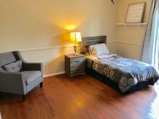 A simple bedroom with a single bed, nightstand and lamp, an upholstered armchair, and wood flooring.