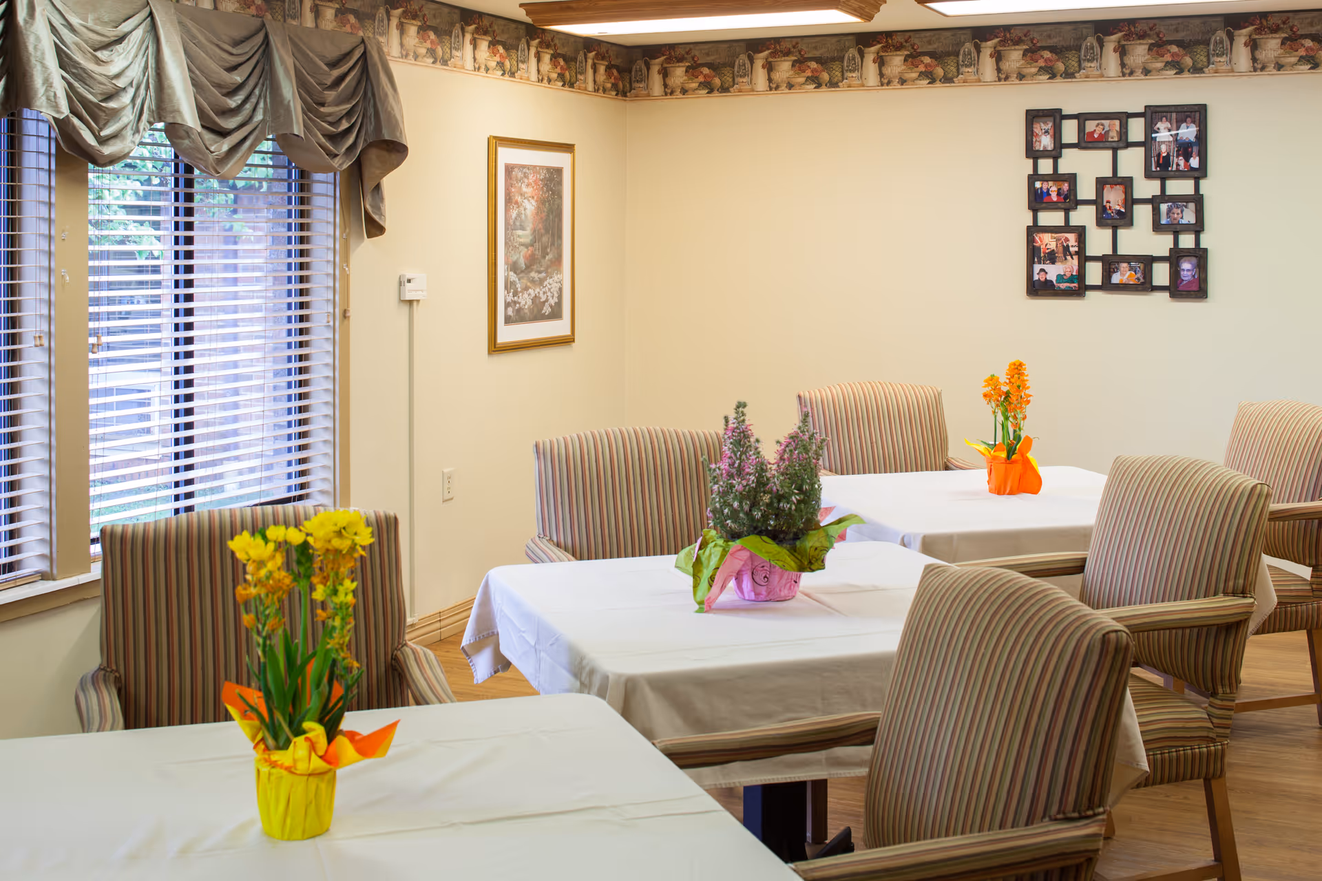 Bright dining room with tables covered in white tablecloths, striped chairs, and small potted floral centerpieces.