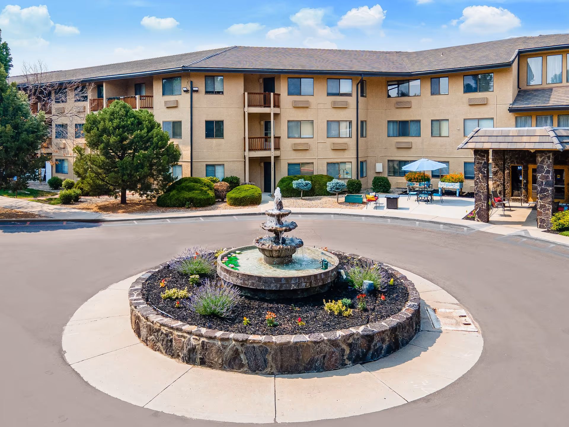 Exterior view of a three-story senior living facility with beige walls and multiple windows. In front of the building is a circular driveway with a stone fountain in the center surrounded by flowers and greenery. There are outdoor seating areas with tables, chairs, and umbrellas near the entrance. The sky is blue with scattered clouds.