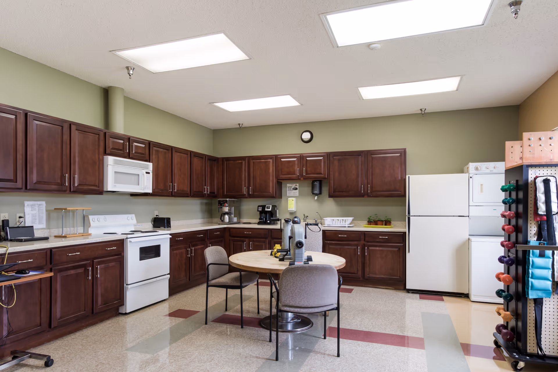 A spacious kitchen area with dark wooden cabinets, white appliances including a stove, microwave, refrigerator, and stacked washer and dryer. There is a round table with two chairs in the center of the room, and a rack holding colorful dumbbells and exercise equipment on the right side. The walls are painted light green and the ceiling has several rectangular fluorescent lights.