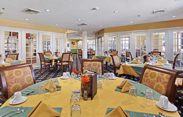 A spacious dining room with multiple tables set for a meal. Each table has yellow tablecloths with green placemats, folded yellow napkins, upside-down glasses, white coffee cups, and silverware. The chairs have patterned upholstery with warm colors. The room features large windows and glass-paneled doors, allowing natural light to fill the space.