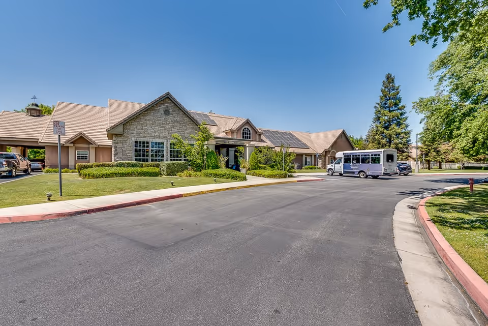 Exterior view of Riverstone Terrace Senior Living facility showing a single-story building with stone and stucco facade, solar panels on the roof, a driveway with a small shuttle bus parked, surrounded by green lawns and trees under a clear blue sky.